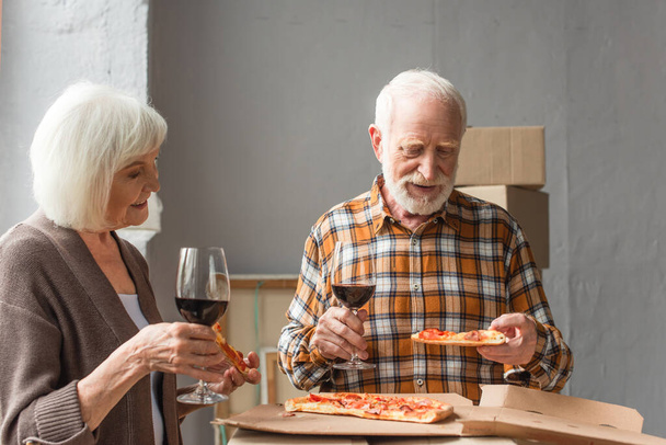 seniorenpaar met stukken pizza en een glas wijn in nieuw huis - Foto, afbeelding