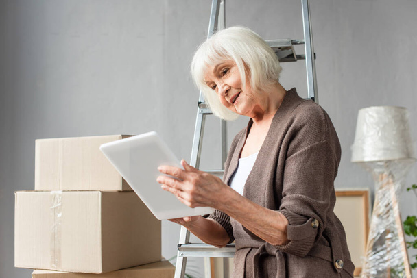 senior woman sitting on ladder and using digital tablet with cardboard boxes on background, moving concept - Photo, Image