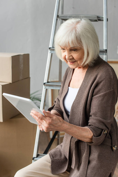 starší žena sedící na žebříku a používající digitální tablet s lepenkovými krabicemi na pozadí, pohybující se koncept - Fotografie, Obrázek