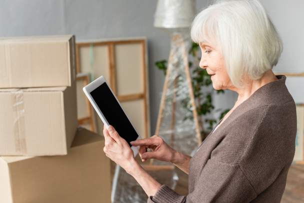 πλαϊνή όψη της ηλικιωμένης γυναίκας κρατώντας ψηφιακό δισκίο με λευκή οθόνη με κουτιά από χαρτόνι στο παρασκήνιο, κινούμενη έννοια - Φωτογραφία, εικόνα