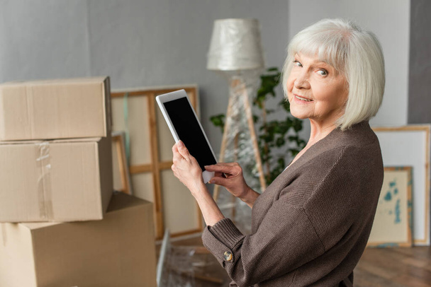 starší žena držící digitální tablet s prázdnou obrazovkou a ohlížející se zpět s kartónovými krabicemi na pozadí, pohybující se koncept - Fotografie, Obrázek