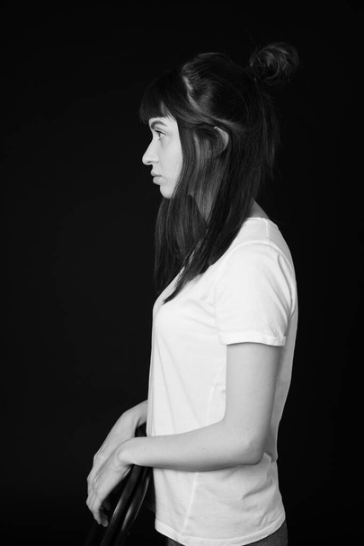 Sideview studio portrait of a pretty brunette woman in a white blank t-shirt, against a plain black background, looking to the side - Photo, image
