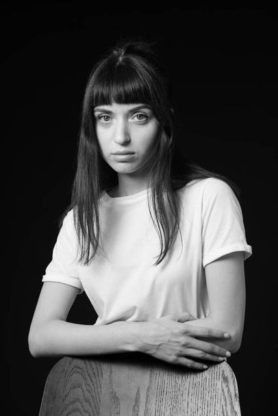 Studio portrait of a pretty brunette woman in a white blank t-shirt, standing behind the chair leaning on it, with crossed arms, against a plain black background, looking at the camera - Фото, зображення