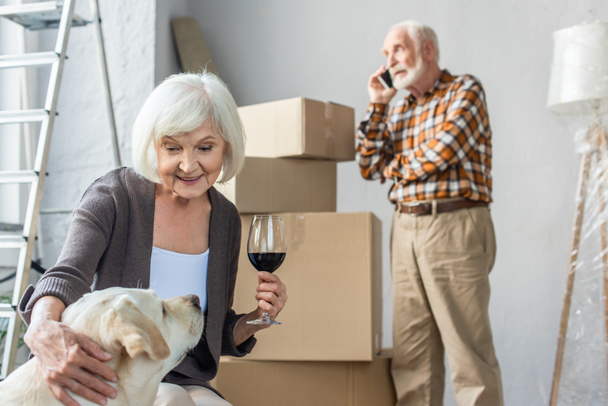 senior woman cuddling dog and holding glass of wine while man talking on phone - Photo, Image