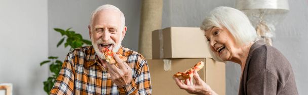 panoramic shot of laughing senior couple eating pizza in new house and cardboard boxes on background - Fotografie, Obrázek