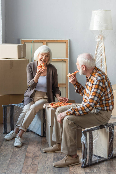 pełny widok długości śmiech senior para jedzenia pizzy w nowym domu i kartonowe pudełka na tle - Zdjęcie, obraz