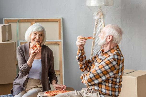 laughing senior couple eating pizza in new house and cardboard boxes on background - Foto, Bild