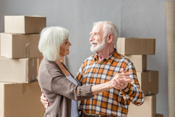 cheerful senior couple dancing in new house with cardboard boxes on background - Foto, imagen