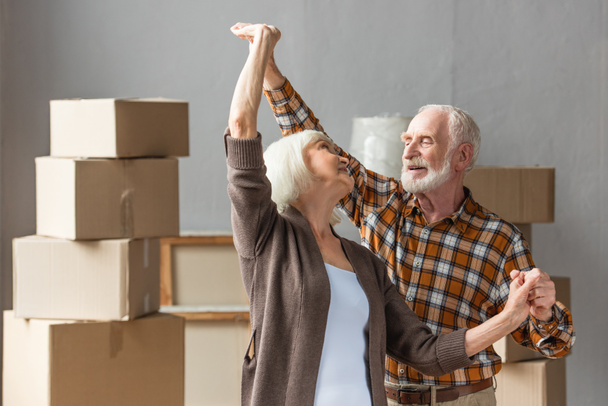 joyeux couple de personnes âgées dansant dans une nouvelle maison avec des boîtes en carton sur fond - Photo, image
