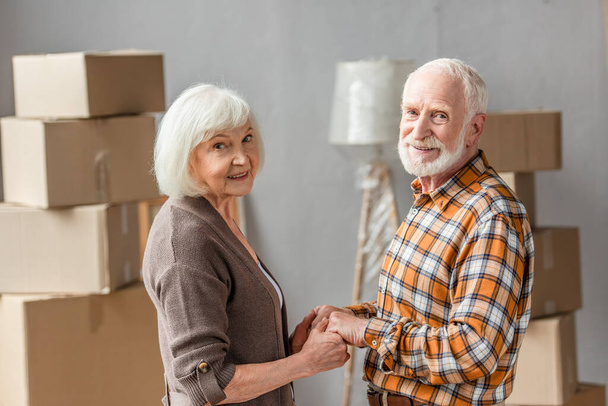 senior couple holding hands and looking at camera in new house, moving concept - Photo, Image