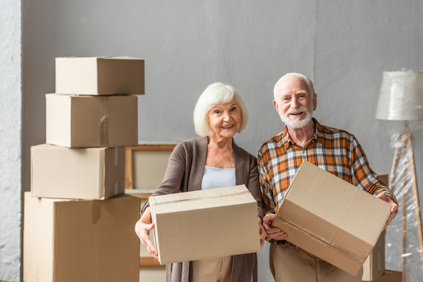 pareja de ancianos sosteniendo cajas de cartón y mirando a la cámara en la nueva casa, concepto en movimiento - Foto, imagen