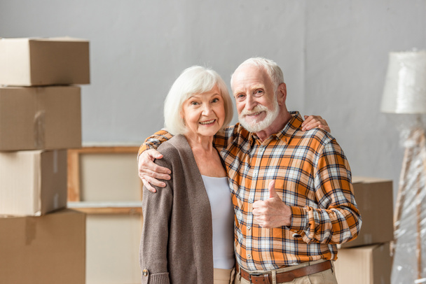 senior couple embracing and man showing thump up in new house, moving concept - Photo, Image
