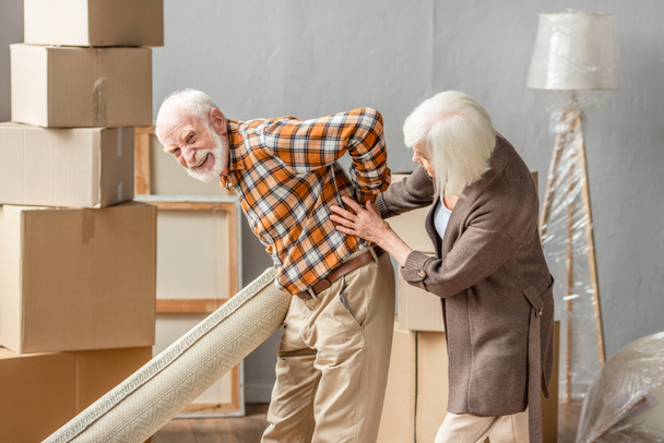 hombre mayor que sufre de dolor de espalda y la celebración de la alfombra, mientras que la esposa tratando de ayudar - Foto, imagen