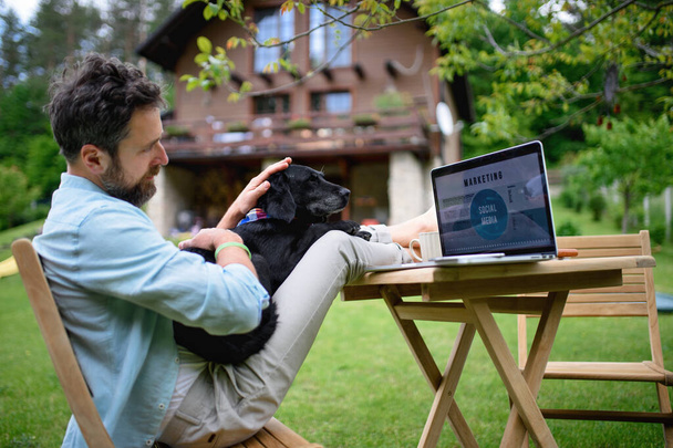 Mature man with laptop and dog working outdoors in garden, home office concept. - Fotó, kép