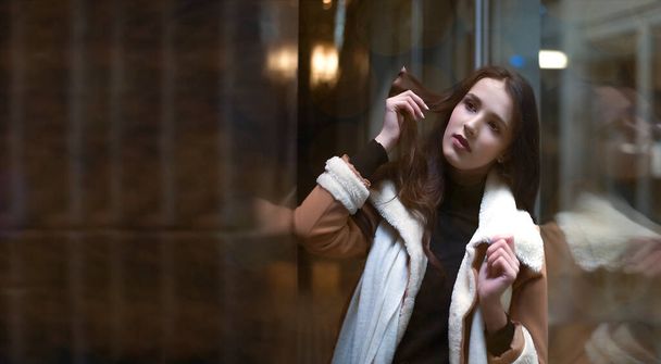portrait of latin hispanic young adult woman stand near big shop window. Brunette asian female girl wearing luxury coat with white fur. Evening shopping. reflection in showcase. dreamy face. Night scene - Foto, Imagem