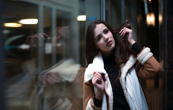 portrait of latin hispanic young adult woman stand near big shop window. Brunette asian female girl wearing luxury coat with white fur. Evening shopping. reflection in showcase. dreamy face. Night scene - Фото, изображение