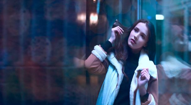 portrait of latin hispanic young adult woman stand near big shop window. Brunette asian female girl wearing luxury coat with white fur. Evening shopping. reflection in showcase. dreamy face. Night scene - Foto, Imagen