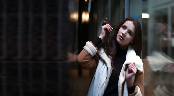 portrait of latin hispanic young adult woman stand near big shop window. Brunette asian female girl wearing luxury coat with white fur. Evening shopping. reflection in showcase. dreamy face. Night scene - Fotó, kép