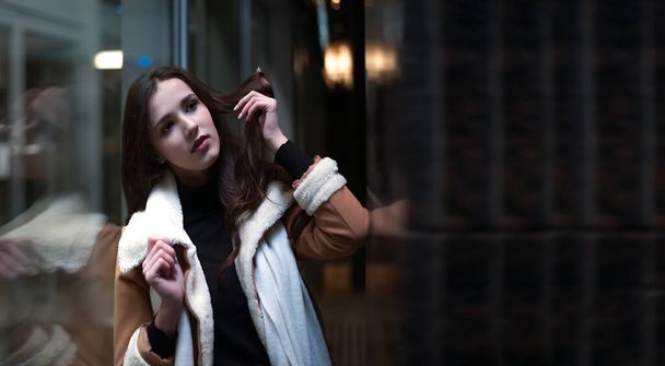 portrait of latin hispanic young adult woman stand near big shop window. Brunette asian female girl wearing luxury coat with white fur. Evening shopping. reflection in showcase. dreamy face. Night scene - Foto, immagini