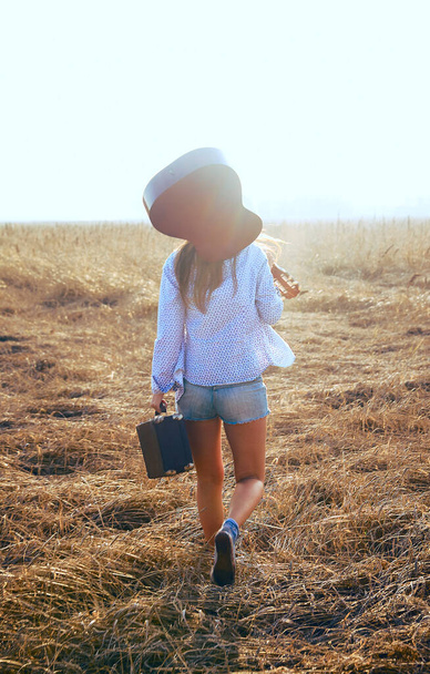 hippie girl walking in wheat field with acoustic Guitar. old retro suitcase in hand. back view. - Fotó, kép