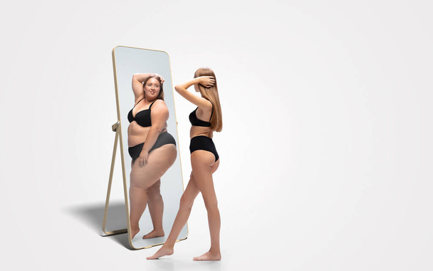Young slim woman looking at fat girl in mirrors reflection on white background - Fotó, kép