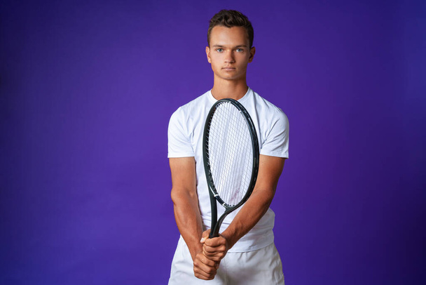 Caucasian young man tennis player posing with tennis racket against purple background - Fotó, kép