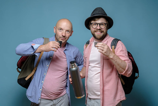 Two happy male tourists with city backpacks drink a hot drink from a thermos and stare into the camera. Blue background. - Fotó, kép