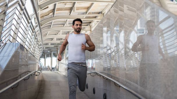 Knappe Latijns-Amerikaanse baard jongeman in sportkleding loopt op skywalk brug in de stad of stad. Bodybuilding en gezonde levensstijl concept. Sportieve man joggen oefening in de stad. - Foto, afbeelding