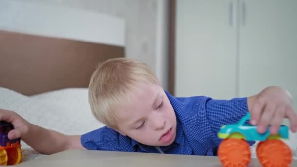 παιδική ηλικία με αναπηρίες, πορτρέτο του αγοριού με σύνδρομο Down παίζει με το αυτοκίνητο παιχνίδια που βρίσκονται στο κρεβάτι στο δωμάτιο στο σπίτι - Πλάνα, βίντεο