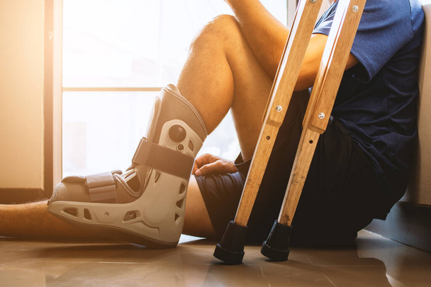 Verletzter Mann gebrochen Knöchel trägt Knöchelstütze sitzt im Haus mit Soft-Fokus und über Licht im Hintergrund - Foto, Bild