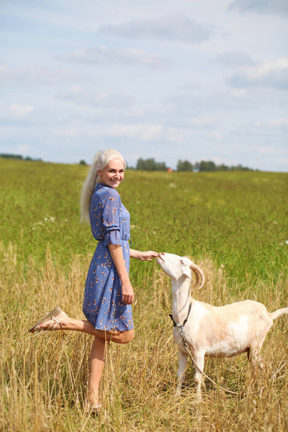 Portrait of a young blonde girl with a goat in a wheat field - Photo, Image
