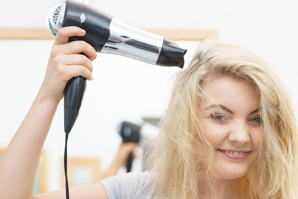 Positive woman using hair dryer on her blonde hairdo. Haircare, hairstyling concept. - Photo, image