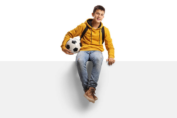 Teenager boy holding a soccer ball and sitting on a white panel board isolated on white background - Foto, Imagen