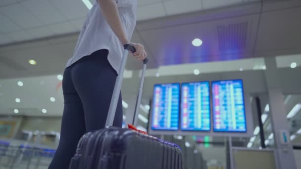 Parte inferiore del corpo della giovane donna in piedi, guardare la schermata orario del volo con i bagagli in viaggio, al terminal dell'aeroporto, in movimento, servizio di biglietteria delle compagnie aeree, donna con valigia - Filmati, video