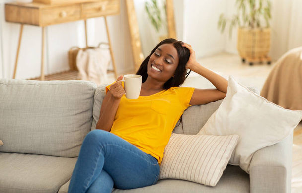 Pretty young black lady resting on sofa with cup of hot drink at home - 写真・画像