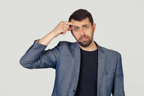 Young businessman man with a beard in a jacket, pointing to an unhappy pimple on his forehead, an ugly acne infection. Acne and skin problems. Portrait of a man on a gray background. - Photo, image