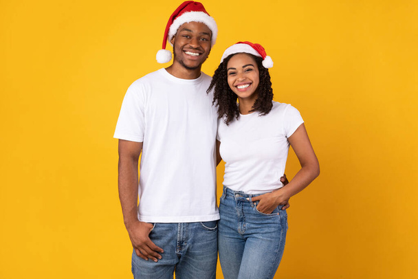 Vreugdevol Afrikaans paar in Santa Kerstmis hoeden poseren, gele achtergrond - Foto, afbeelding