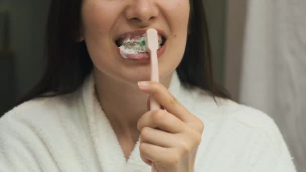 Spiegelung im Spiegel einer jungen Frau im Bademantel, die ihre Zähne mit Zahnspangen mit Zahnbürste putzt - Filmmaterial, Video