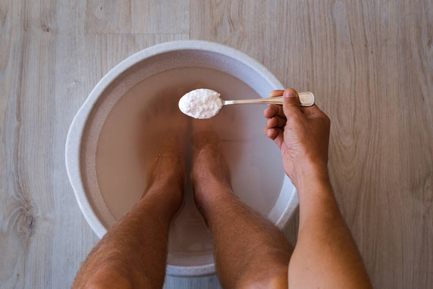 De man deed een lepel zuiveringszout in bad met heet water voor zijn voeten. Zelfgemaakte bad weken voor droge voeten huid - Foto, afbeelding