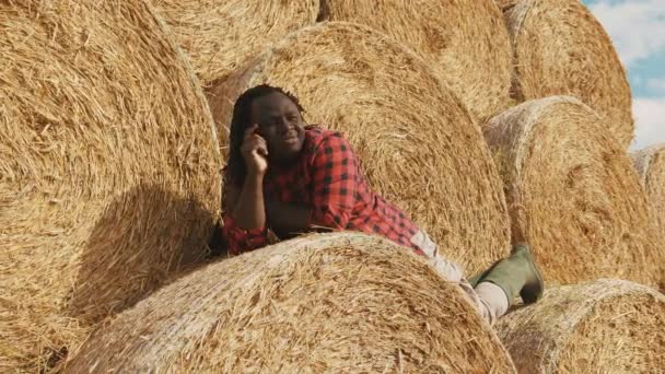 Νεαρός ευτυχισμένος Αφρικανός αγρότης ξαπλωμένος στη στοίβα από ρολά σανού και ξεκουράζεται - Πλάνα, βίντεο
