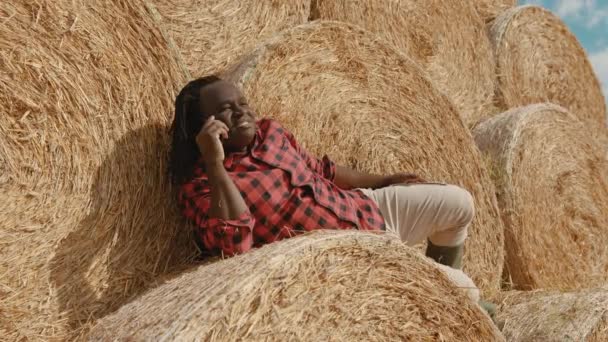 Joven agricultor africano feliz acostado en la pila de rollos de heno, descansando y hablando por teléfono - Imágenes, Vídeo