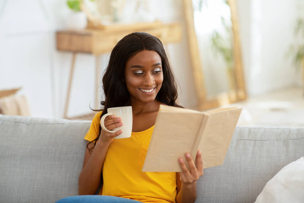 Залишайтеся на дозвіллі додому. Прекрасна молода чорна жінка читає книгу з чашкою гарячого напою у вітальні
 - Фото, зображення