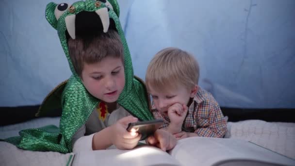 Der große Bruder im Drachenkostüm spielt mit einem Jüngeren, Kinder benutzen ein Smartphone und lesen im heimischen Zelt Bücher - Filmmaterial, Video