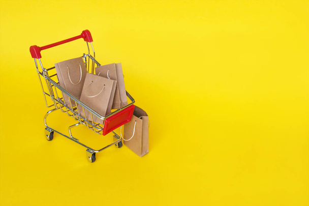 Mini carrello della spesa con piccole borse artigianali sullo sfondo giallo. Il concetto di shopping nei negozi e online. - Foto, immagini