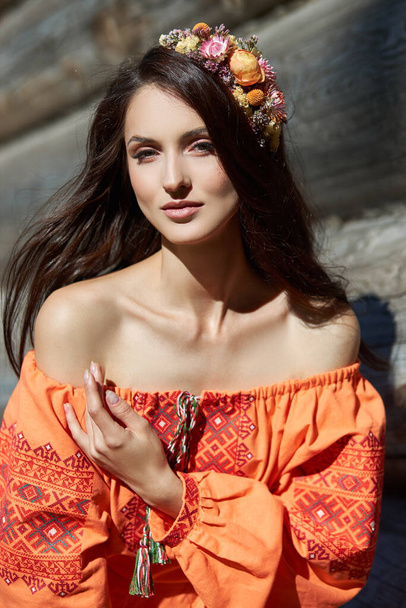 Beautiful Slavic woman in an orange ethnic dress and a wreath of flowers on her head. Beautiful natural makeup. Portrait of a Russian girl - Foto, Imagem