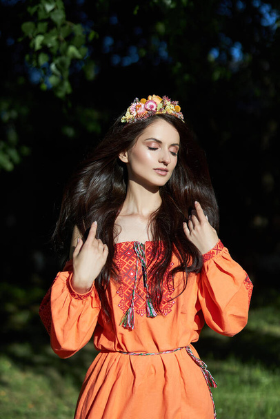 Beautiful Slavic woman in an orange ethnic dress and a wreath of flowers on her head. Beautiful natural makeup. Portrait of a Russian girl - Фото, зображення