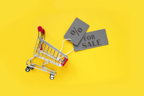 Mini carrito de compras con etiquetas grises con una inscripción EN VENTA sobre un fondo amarillo. El concepto de compras en tiendas y en línea. - Foto, imagen