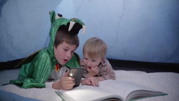 older brother in a dragon costume plays with a younger, children use a mobilephone with flashlight and read book while lying in tent at home - Footage, Video