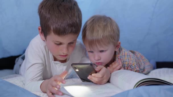 older brother reads fairy tales for younger and uses a phone with flashlight lying in a tent at home during vacation - Footage, Video