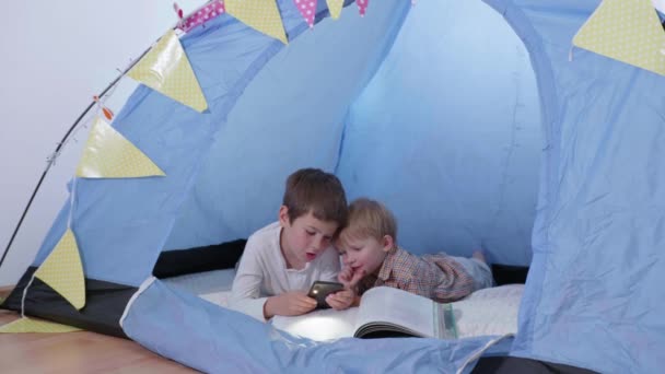 καλύτεροι παιδικοί φίλοι, δύο αδέλφια διαβάζουν ένα βιβλίο και χρησιμοποιούν ένα τηλέφωνο με φακό ξαπλωμένο σε μια σκηνή στο σπίτι - Πλάνα, βίντεο